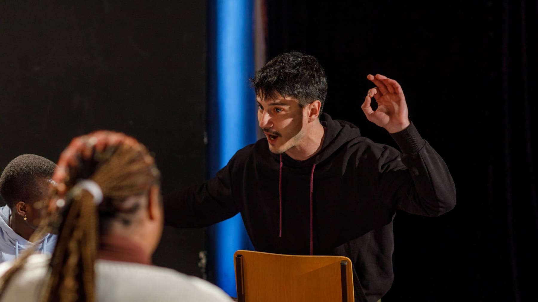 Un improvisateur en sweat noir s'adresse à ses partenaires pendant une scène, sur un plateau sombre